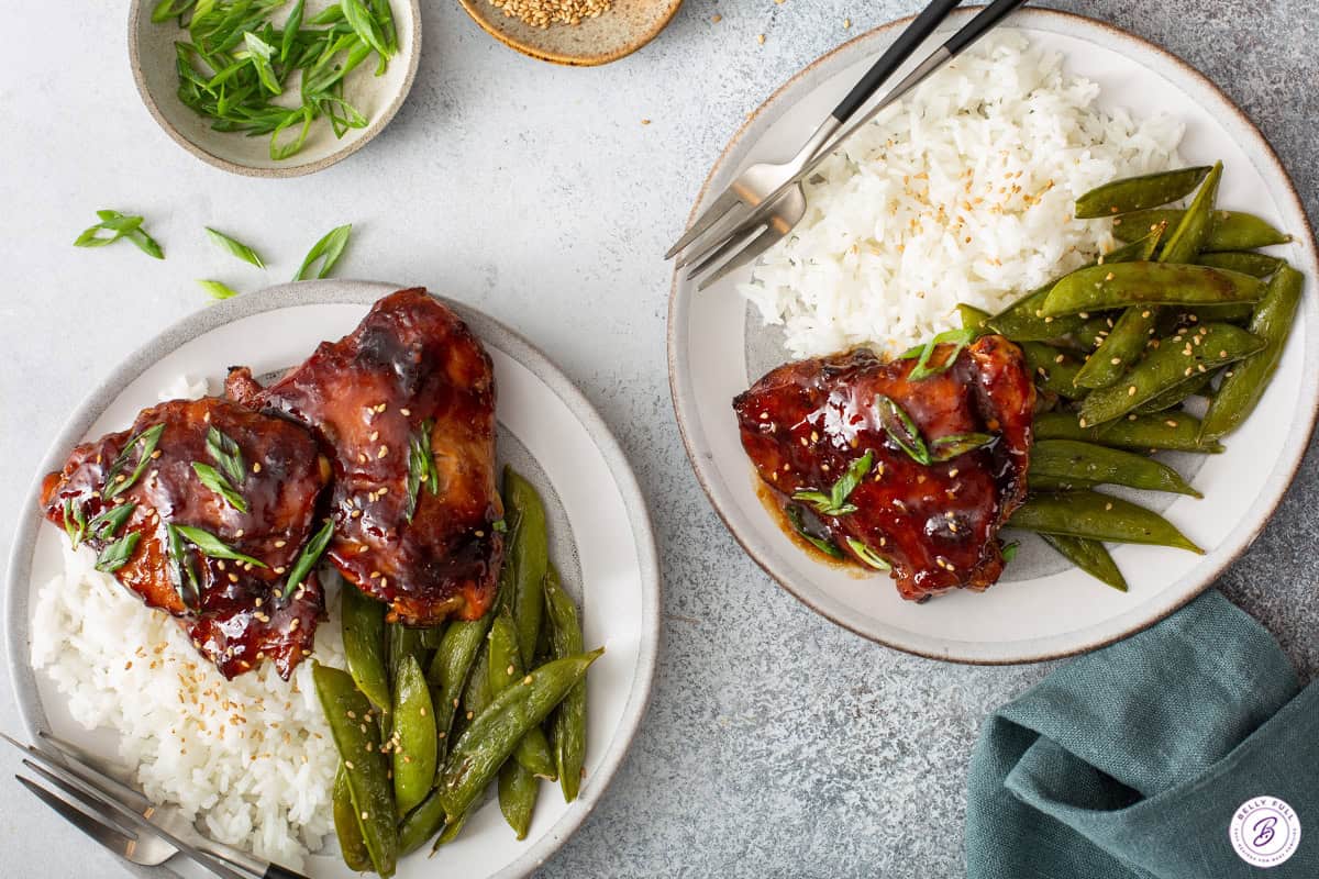 Overhead shot of two plates of teriyaki chicken served with white rice and roasted snap peas, chopsticks and a fork resting alongside.