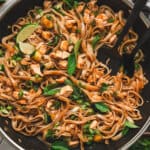 Overhead shot of Thai drunken noodles in a stainless steel wok, wide rice noodles tossed with chicken, zucchini, scallions, and Thai basil, with lime wedges tucked among the noodles.