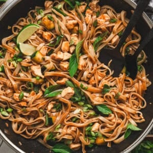 Overhead shot of Thai drunken noodles in a stainless steel wok, wide rice noodles tossed with chicken, zucchini, scallions, and Thai basil, with lime wedges tucked among the noodles.