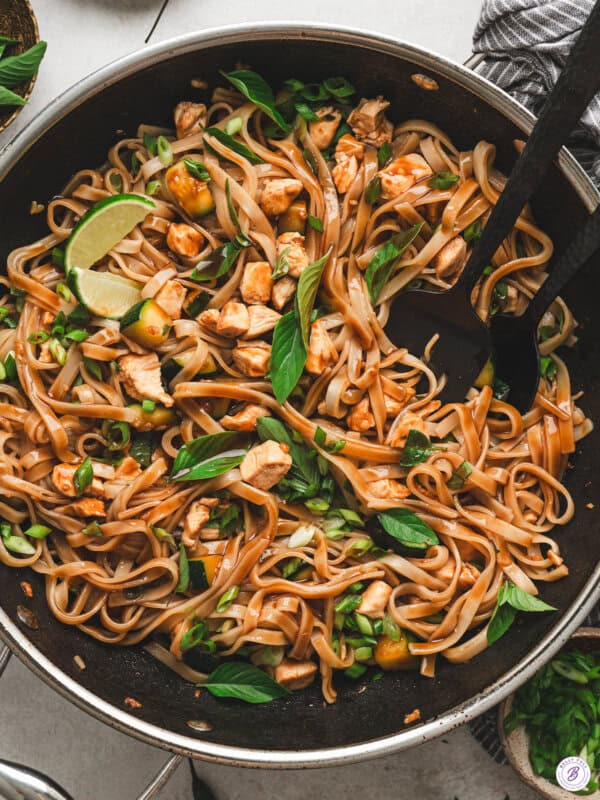 Overhead shot of Thai drunken noodles in a stainless steel wok, wide rice noodles tossed with chicken, zucchini, scallions, and Thai basil, with lime wedges tucked among the noodles.
