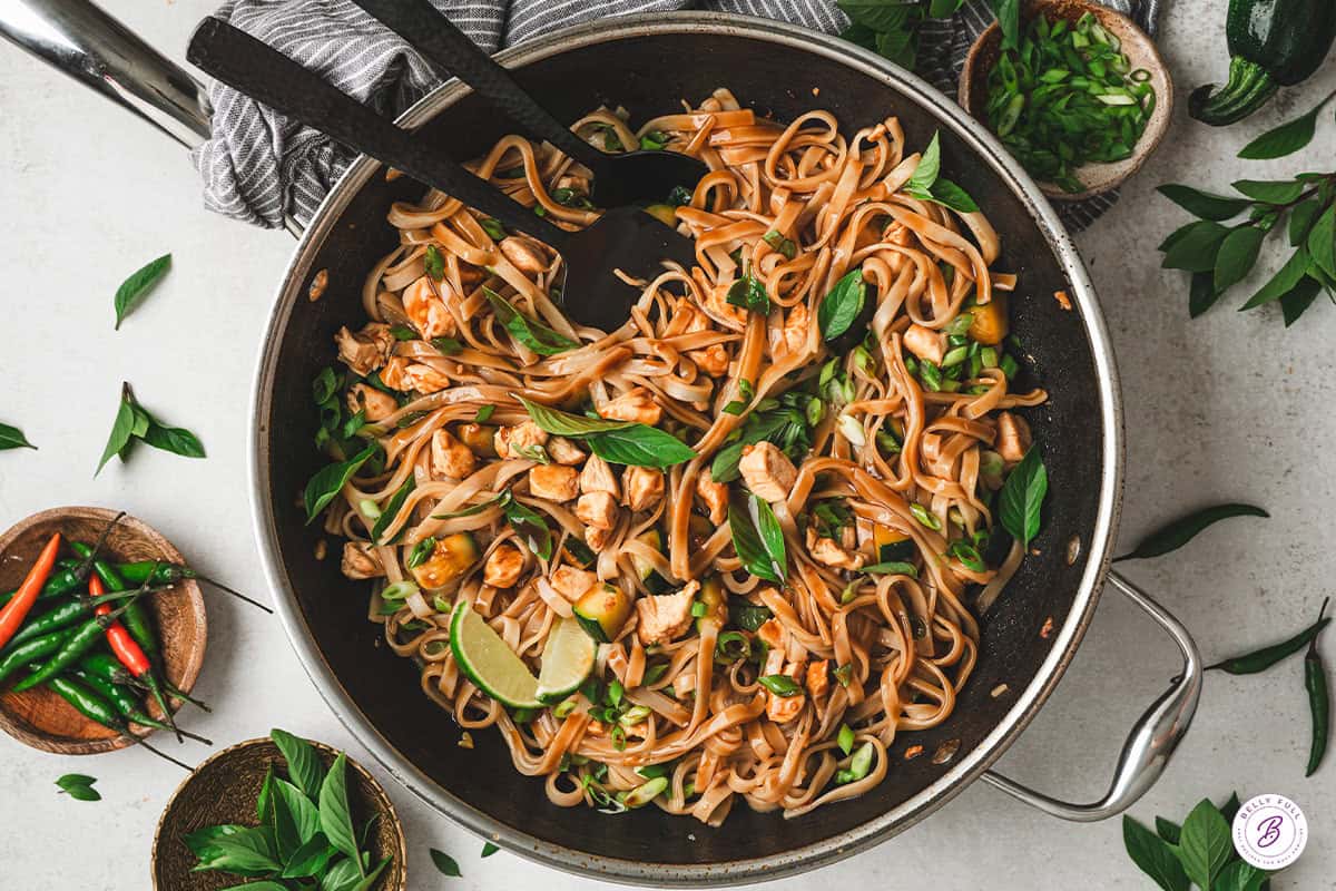Overhead shot of finished drunken noodles in a wok with serving spoons, surrounded by fresh Thai basil, red and green Thai chilies, and lime wedges.