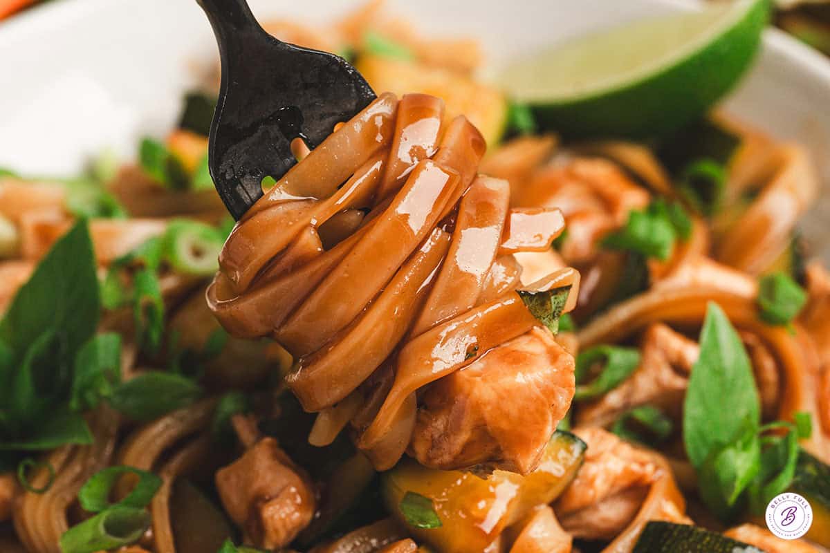 Close up of a forkful of glossy drunken noodles lifted from the plate, with a piece of chicken and fresh Thai basil visible.