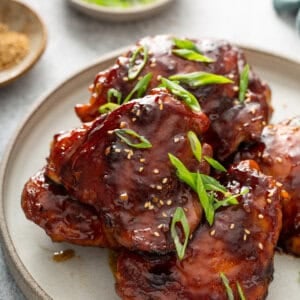 Close-up angled shot of glazed teriyaki chicken thighs piled on a plate, the sauce deep mahogany and glossy, topped with sliced scallions and sesame seeds, with small bowls of scallions and sesame seeds in the background.