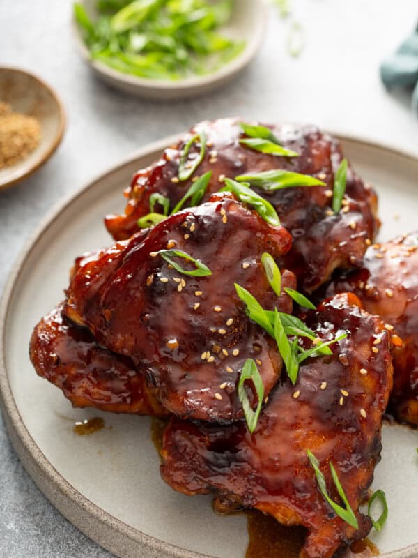 Close-up angled shot of glazed teriyaki chicken thighs piled on a plate, the sauce deep mahogany and glossy, topped with sliced scallions and sesame seeds, with small bowls of scallions and sesame seeds in the background.