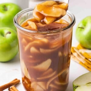 Spoon lifting a glossy apple slice from a jar of homemade apple pie filling, surrounded by whole Granny Smith apples and cinnamon sticks.