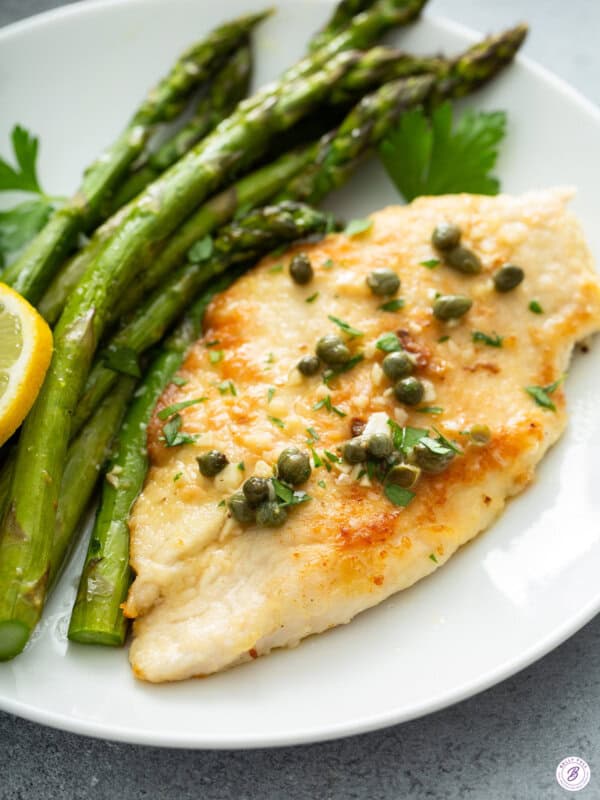 Close-up angled shot of pan-seared chicken piccata topped with capers, minced garlic, and chopped parsley, plated next to roasted asparagus spears and a lemon half, with a parsley leaf garnish in the background.