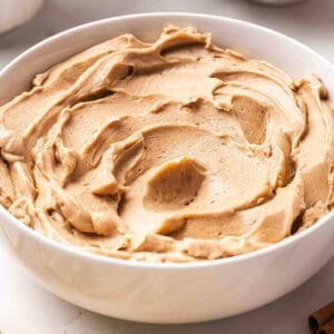 Close-up overhead shot of whipped Texas Roadhouse cinnamon butter swirled in a white bowl, with a scalloped dish of honey blurred in the background and a whole cinnamon stick resting nearby.