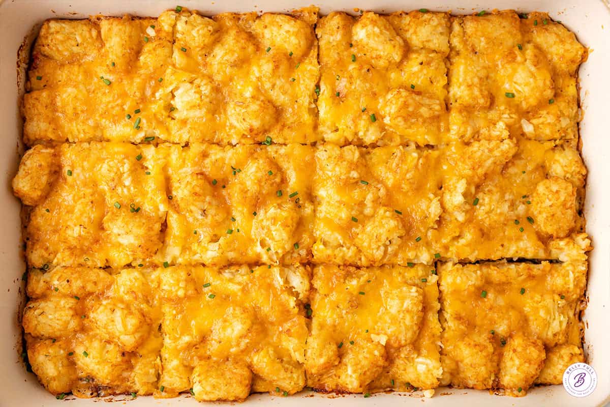Close-up of tater tot breakfast casserole in a white baking dish, cut into squares with melted cheddar cheese and chives