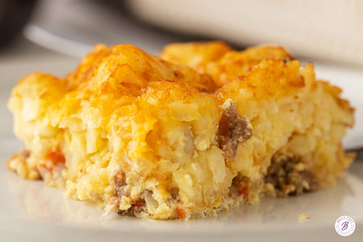 Close-up of a slice of tater tot breakfast casserole on a white plate, showing the tater tot, egg, and sausage layers